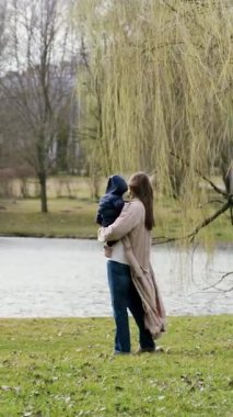 Bir anne, su kenarındaki sakin bir parkta yürürken çocuğunu kucaklıyor. Kamera, neşeli hareketlerini takip ediyor. Aile bağının özünü yakalıyor.