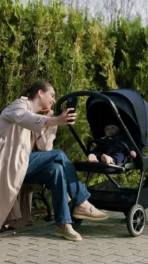 Bej ceketli bir kadın, park yolunda bebek arabasının yanında otururken akıllı telefonuyla neşeli selfie çekiyor, neşeli anı ve çevresini saran yemyeşil manzarayı vurguluyor