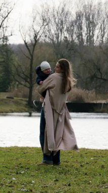 Dalgalı ceketli kadın çocuğu sakin göl kenarında tutarken, sevgi ve bağlantıyı vurguluyor, kamera da bu hareketi hareketli bir hareketle takip ediyor.