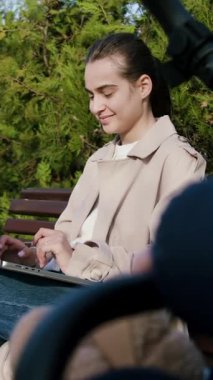 Bej renkli trençkotlu bir kadın, dizüstü bilgisayarıyla bir park bankında meşgul, yemyeşil, yoğun konsantrasyon ve sükunet gösteren kamerayla çevrili,