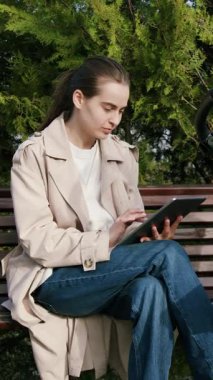 Bej renkli trençkotlu kadın, park bankında tabletle meşgul, bebek arabasının yanında, modern uzaktan çalışmayı gösteren, kamera onu dolly hareketleriyle takip ediyor.