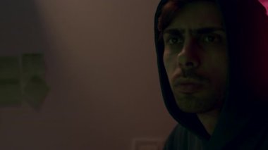 Koyu renk kapüşonlu genç adam derin bir şekilde yansıyor. Düşünceli ifadesini ve odanın ışıklandırmasını gösteren kamera görüntüleriyle duygusal rezonansı arttırıyor.