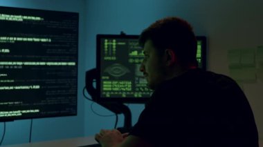 Erkek yapay zeka mühendisi düşük ışık ortamında çift monitörlerde gösterilen kodlarla derinden ilgileniyor. Analitik yeteneklerini ve teknolojinin atmosferini vurguluyor.