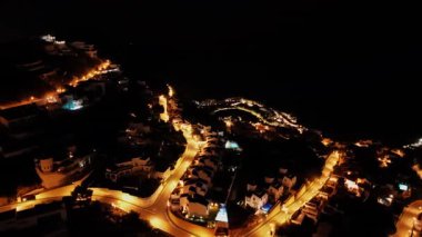 Hava perspektifi, geceleri aydınlatılmış bir kıyı köyünü yakalıyor, dolambaçlı yolları ve parlayan evleri vurguluyor, giderek uzaklaşıp manzarayı gözler önüne seriyor.