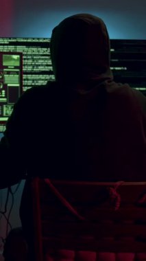 Kapüşonlu eşofman üstü giyen bir birey karmaşık verileri ve canlı grafikleri gösteren birden fazla ekrana odaklanır, yavaş yavaş çalışma alanı kurulumunu ortaya çıkarmak için uzaklaşır