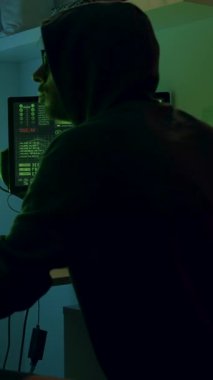 Kapüşonlu bir kişi verilerle dolu bilgisayar ekranlarına odaklanıyor, yüksek teknolojili bir çalışma alanı sergiliyor, kameralı bebek olay yerini yakalamak için sorunsuzca içeri giriyor