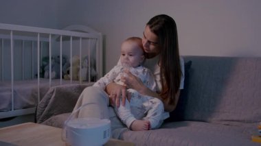 Anne, bebeğini sıcak ve davetkar bir ortamda solunum terapisi sırasında sakinleştirir. Sevgi etkileşimini vurgular. Duygusal bağlantıyı vurgulamak için kamera tavaları.