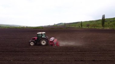 Elinde bir tohum matkabı olan kırmızı bir traktör geniş bir araziyi verimli bir şekilde tarıyor, zengin, karanlık toprakta düzgün çizgiler oluşturuyor, bu da tarımsal makinelerin iş başında olduğunu gösteriyor.