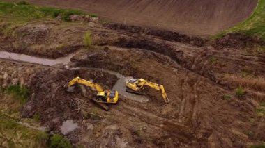 İki kazıcı, bir su kanalı oluştururken toprak ve enkazı kaldırmak için işbirliği yaparak, bir inşaat alanında ağır makineler gösteriyor