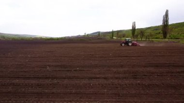 Bir kırmızı traktör geniş bir tarlayı sürmeye çalışır, toprağı döndürür ve çizgiler oluşturur toz bulutları oluşurken, hareket halindeki tarımsal süreçleri vurgular.
