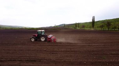 Kırmızı bir traktör yeni sürülmüş bir tarlaya tohum eker, toz bulutu yaratır ve açık tarım arazilerindeki tarım sürecini gözler önüne serer.