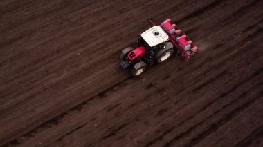 Elinde tohum matkabı olan kırmızı bir traktör, tohum ekerek tarımsal makineleri ve tarım tekniklerini ön plana çıkaran, zengin, karanlık toprağa sürekli olarak ilerler.