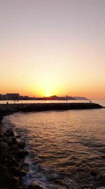 Gün batımında çarpıcı bir kıyı şeridinin büyüleyici hava manzarası, su yüzeyindeki canlı renkleri vurguluyor, pürüzsüz bir kamera zoom ile sakin atmosferi güçlendiriyor.