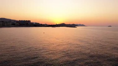Sakin kıyı günbatımı sahnesi, sakin sular sıcak renkleri yansıtıyor, kamera zarafetle hareket ediyor, akşam gökyüzünün ve manzaranın dingin güzelliğini gözler önüne seriyor.