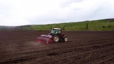 Tohum ekicisi ile donatılmış kırmızı bir traktör tarladan geçerek toprağa tohum dağıtarak tarım ortamında bir toz bulutu oluşturur.