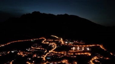 Hava perspektifi, alacakaranlıkta aydınlatılmış bir dağ kasabasını yakalıyor, dolambaçlı sokakları ve parlayan ışıkları aydınlatıyor, pürüzsüz bir kamera ile geniş manzarayı gözler önüne seriyor