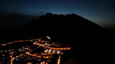 Hava perspektifi, bir dağın altındaki ışıklarla parlayan bir köyü ortaya çıkarıyor, dingin geçişi geceden alacakaranlığa yansıtıyor, kamera peyzaj boyunca sorunsuzca hareket ediyor.
