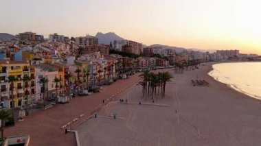 Hava perspektifi, gün batımında canlı bir kıyı kasabasını yakalar, plaj boyunca renkli mimari ve palmiye ağaçlarını aydınlatır, sakin sahneyi güzelleştiren yumuşak bir kamera tavasıyla