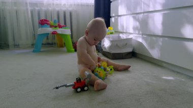 Genç bir çocuk çeşitli oyuncaklarla etkileşime girer, yumuşak bir halıda otururken şekilleri ve renkleri inceler, parlak kapalı bir alanda neşeli elementlerle çevrilidir.