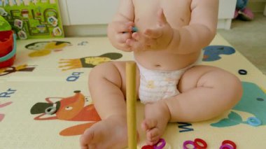 Bir bebek ahşap bir direğe renkli yüzükler yığar, neşeli tasarımlarla dolu canlı bir oyun minderinde otururken iyi motor becerileri ve merakını gösterir.
