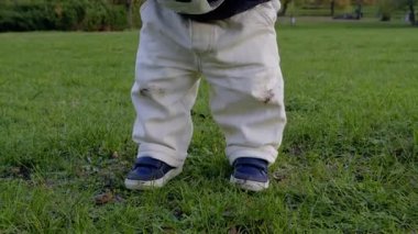 Beyaz pantolon ve mavi ayakkabı giymiş bir çocuk yeşil çimlerin üzerinde duruyor, futbol topunu tekmelemeye hazır, açık havada oyuncu bir enerji ve beklenti sergiliyor.