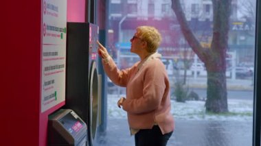 Pembe kazaklı yaşlı bir kadın çamaşırhane makinesiyle takılıyor. Çamaşır talimatlarını açık hava manzaralı, aydınlık ve çağdaş bir mekanda inceliyor.