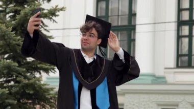 Mezuniyet kıyafetli bir erkek öğrenci, açık hava fotoğraflarına poz veriyor, bir üniversite binasının önünde şapkasını ayarlıyor ve olay yerini çevreleyen ağaçlarla çevrili.