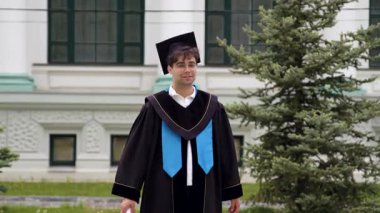 Siyahi mezuniyet kıyafetleri içinde erkek mezun açık havada duruyor, üniversite binasının önünde diplomasını tutuyor ve arka planda yeşillik var.