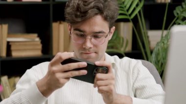 Gözlüklü genç adam rahat bir atmosfer için etrafı kitaplık ve yeşilliklerle çevrili akıllı telefonunu rahat bir çalışma odasında görüyor.
