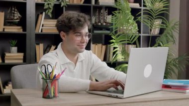 Kıvırcık saçlı erkek öğrenci, çağdaş çalışma alanındaki kitaplar ve yeşil bitkilerle çevrili ahşap bir masadaki dizüstü bilgisayarda yazmaya odaklanıyor.