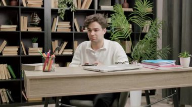 Beyaz tişörtlü beyaz bir adam oyun kontrolörüyle bir masada oturur, kitap rafları ve rahat çalışma ortamındaki kapalı bitkilerle çevrili.