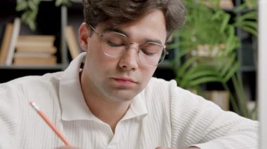 Beyaz kazaklı erkek öğrenci, çalışma odasında kitaplarla ve ev bitkileriyle çevrili bir masada kalemle notlar yazmaya odaklanıyor.