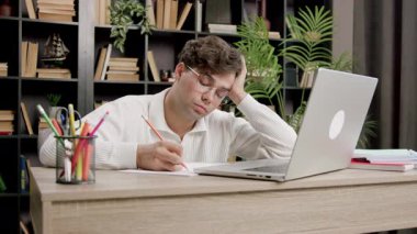 Gözlüklü erkek öğrenci bir masaya notlar yazıyor, bir dizüstü bilgisayara odaklanıyor, kitaplar ve bitkiler sıcak bir çalışma ortamı yaratıyor.