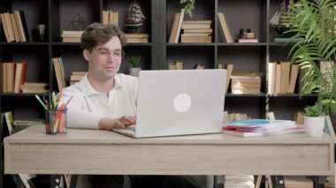 Beyaz kazaklı genç adam, kitaplarla ve dekoratif eşyalarla dolu ahşap bir masada dizüstü bilgisayar kullanıyor ve rahat bir çalışma ortamı yaratıyor.