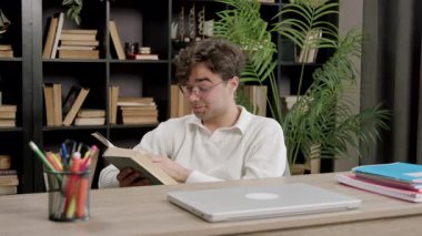 Gözlüklü genç adam kitap rafları ve kırtasiyelerle çevrili bir masada kitap okuyor ve sıcak ve davetkar bir çalışma ortamı yaratıyor.