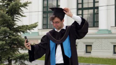 Siyah cüppeli ve mavi elbiseli bir erkek mezun şapkayı ayarlarken ve üniversite binasının önünde yeşil bir selfie çekerken gülümsüyor.