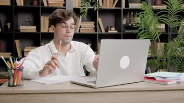 Gözlüklü öğrenci bir masada oturur, elinde bir kalem, etrafında kitap ve kırtasiye malzemesi tutarken dizüstü bilgisayarına konsantre olur.