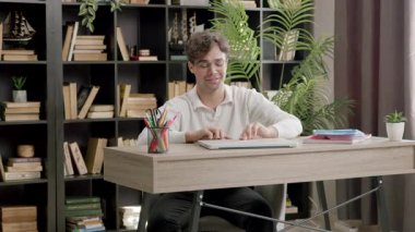 Bir masadaki öğrenci dizüstü bilgisayarla meşgul oluyor, kitaplıklar ve bitkilerle sıcak bir çalışma ortamı yaratıyor.