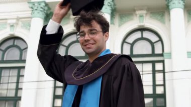 Mezuniyet kıyafetli bir erkek öğrenci, üniversitenin önünde şapkasını neşeyle sallıyor ve arkasındaki binanın etkileyici mimarisini gözler önüne seriyor.