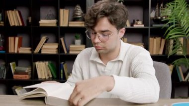 Kıvırcık saçlı genç adam bir masada oturuyor, bir kitabı karıştırıyor, kitaplarla çevrili iken hayal kırıklığını ifade ediyor.