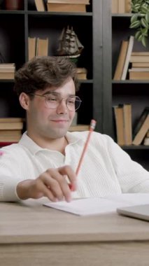 Gözlüklü bir erkek öğrenci, elinde bir kalem, elinde bir dizüstü bilgisayarla rahat bir çalışma ortamında kitap ve kırtasiye malzemeleriyle masada oturuyor.