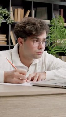 Kıvırcık saçlı öğrenci, kitaplarla dolu samimi bir çalışma odasında laptopuyla ve kırtasiyesiyle özenle masasına notlar yazıyor.