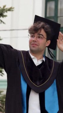 Mezuniyet kıyafetli bir erkek öğrenci, açık hava fotoğraflarına poz veriyor, bir üniversite binasının önünde şapkasını ayarlıyor ve olay yerini çevreleyen ağaçlarla çevrili.