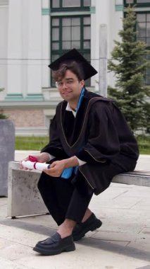 Mezuniyet kıyafetli bir öğrenci, yeşil ve modern mimari ile çevrili bir halde bir bankta oturmuş, başarılarını düşünüyor.