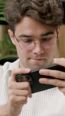 Gözlüklü genç adam rahat bir atmosfer için etrafı kitaplık ve yeşilliklerle çevrili akıllı telefonunu rahat bir çalışma odasında görüyor.