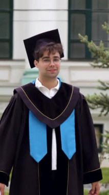Siyahi mezuniyet kıyafetleri içinde erkek mezun açık havada duruyor, üniversite binasının önünde diplomasını tutuyor ve arka planda yeşillik var.