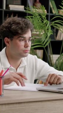 Gözlüklü bir erkek öğrenci, kitaplarla ve bitkilerle dolu rahat bir çalışma alanındaki ahşap bir masada dizüstü bilgisayarına odaklanırken not alıyor.