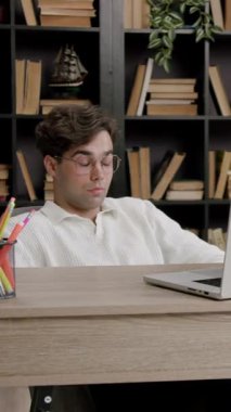 Gözlüklü bir erkek öğrenci, ahşap bir masada dizüstü bilgisayarla, kitap rafları ve rahat bir çalışma odasında dekoratif bitkilerle çalışıyor.