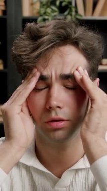 Kahverengi saçlı, beyaz kazaklı bir erkek, arka planda kitaplar ve bitkiler olan bir masada otururken iki eliyle başını tutuyor.