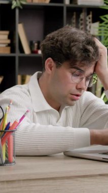 Tahta masadaki dizüstü bilgisayarda oturan beyaz kazaklı genç adam, etrafı kitaplarla ve bitkilerle çevrili, düzenli çalışma alanı olan rahat bir çalışma ortamında.